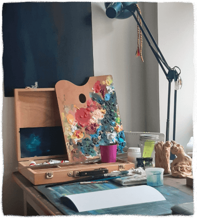 Foto Arbeitsplatz voller verschiedener Materialien Vera Bergs Zeichentisch bestückt mit einem Farbkoffer, einer bunten Farbpalette, Pinseln, Papier und Tonfiguren.