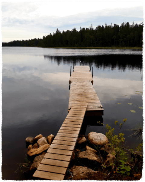 Foto Holzsteg schwedischer See (Tablet-Version) Ein langer Holzsteg in drei Teilen ragt in einen schwedischen See. Die Wasseroberfläche ist klar und beruhigt. im Hintergrund ist Wald, der sich im Wasser spiegelt.