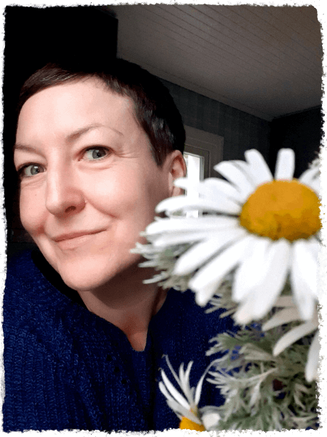 Portrait Vera Berg mit Margarite Vera Berg lächelt fröhlich und schaut seitlich hinter einer Margarite hervor.