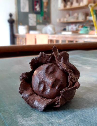 Eine dunkelbraune Blütenkind-Plastik liegt auf dem Arbeitsplatz der Künstlerin. Sie ist komplett fertig gebrannt und glasiert. Die farblose, leicht glänzende Glasur gibt der Plastik einen leichten Schimmer.