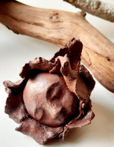 Der dunkle, kahle, runde Kopf eines kleinen Menschen ist umrandet von Blütenblättern im selben Ton. Die Augen sind geschlossen.