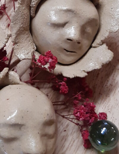 Zwei helle Köpfchen, die im Inneren einer Blüte schlafen, liegen zwischen zart getrockneten pinken Schleierkrautblüten auf einem Holzteller. Eine grün glänzende Murmel findet sich auch inmitten dieser Präsentation.