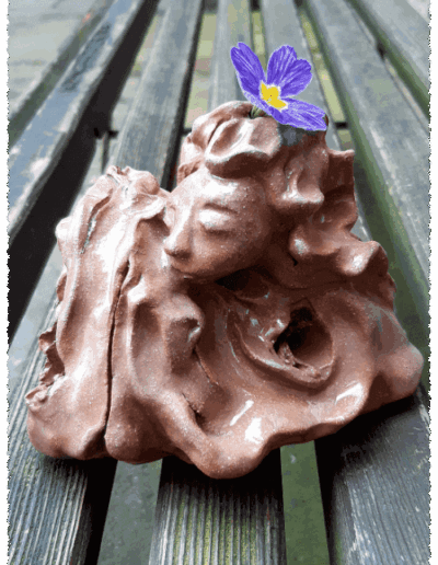 Wie aus einer Art Kleckerburg wachsen zwei Frauen in inniger Umarmung in die Höhe. Nur ihre Köpfe sind klar ausgeformt und dennoch ist erkennbar, wie sie in der Umarmung verschmelzen. Nur eines der beiden Gesichter ist uns zugewandt, die Augen sind geschlossen. Kleine höhlenartige Krater sind in den Körpern erkennbar. Das uns zugewandte Gesicht ist oben auf dem üppigen Haar mit einer echten, blauen Primelblüte geschmückt.