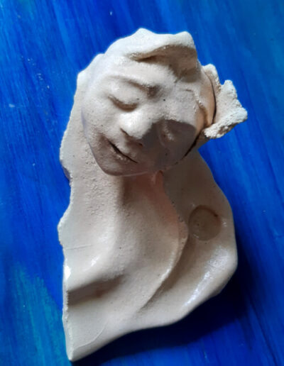 Wie nur eine Hälfte von etwas kommt diese Art Ablagetellerchen aus hellem Ton daher. Es steht auf blauem Grund. Das Gesicht, das ihm entwächst, scheint tief im Träumen versunken und sich aus einer Blüte empor zu winden.