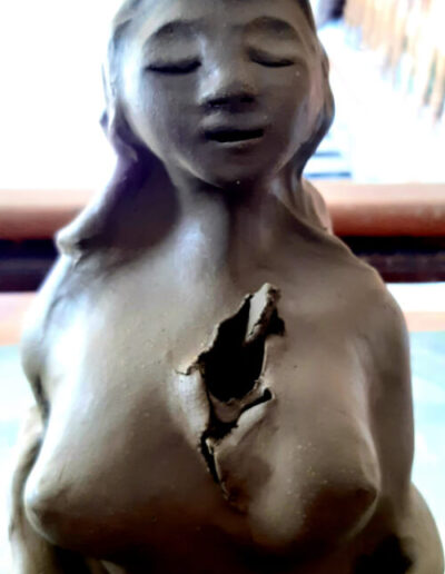 Hier ist im Detail der Oberkörper einer tiefdunklen Mama Cacao zu sehen, die sich aus einer Hälfte der Kakaofrucht erhebt. Ihre Brüste sind üppig, der Kopf erhoben. Sie trägt einen Reif mit Edelstein um den Kopf, die Haare fallen auf ihren Rücken. Den Ausdruck des Gesichts könnte man als „wissend, in sich ruhend“ bezeichnen. Die Arme sind eng am Oberkörper und verschwinden ab den Ellbogen in der Kakaofrucht. Über dem Herzen ist eine offene Wunde, ein Krater zu sehen. Alles sonst an ihr erscheint rund und glatt.