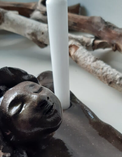 Hier ist eine Detailaufnahme einer dunkelbraunen Ritualschale zu sehen - in Form eines Gesichts mit geschlossenen Augen und einer weißen Kerze daneben.