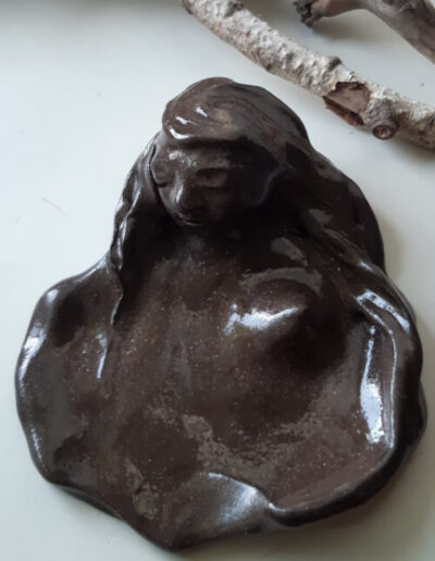 Dieser Teller steht auf hellem Grund und lässt uns einer Frau beim Baden zusehen. Er glänzt in schokoladigem Kakaobraun, ist flach, organisch geformt und einer der größeren meiner Ritualteller. Der weibliche Kopf am einen Ende ist aufrecht. Ein Tuch um die Stirn hält ihre Haare, die links und rechts in den Rand der Schale einfließen. Eine ihrer Brüste erhebt sich aus dem Schalengrund.