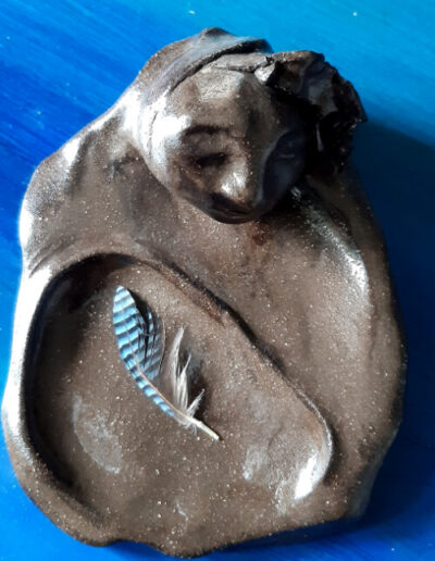 Eine ovale Schale aus schokoladenbraunem Ton steht auf blauem Grund. In diesem Oval befindet sich ein weiteres Oval, leicht vertieft. Darin liegt die kleine, blaue Feder eines Eichelhähers. Am oberen Rand wacht still ein Frauenkopf, der dem Ton entwächst wie einer Blüte.