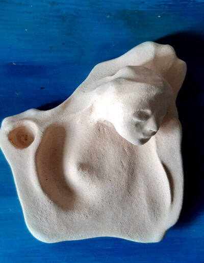 Eine organisch geformte, helle Schale steht auf blauem Grund. Der Blick ist aus der Draufsicht. Im oberen rechten Rand erhebt sich ein Frauenkopf mit geschlossenen Augen,der nach rechts geneigt ist. Haare und Hals fließen in die Begrenzung und in die Schale hinein. Aus dem Fließen entsteht auf der linken Seite die Aussparung für eine kleine Kerze und im Innern der Schale wird der Blick auf die sich leicht, wie aus Wasser erhebende, Brust der Frau gelenkt. Alles fließt um-und miteinander.