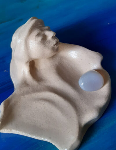 Eine helle Schale, die schmetterlingshaft zu zwei Seiten ausgerichtet ist, steht auf blauem Grund. Mittig zwischen den beiden Flügeln erwächst ein Kopf. Die Augen sind geschlossen. Haare und das Tuch um den Kopf fließen organisch in die Begrenzung der Schale. In einer Schalenhälfte liegt ein bläulich schimmernder Halbedelstein.