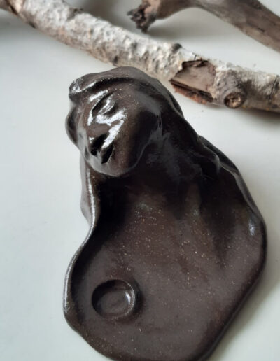 Ein kleiner, dunkelbrauner Ritualteller steht auf hellem Grund. Der träumende Kopf einer Frau mit langem Haar und Tuch umfasst ihn zur Hälfte. Auch hier gibt es die Aussparung für eine kleine Kerze.