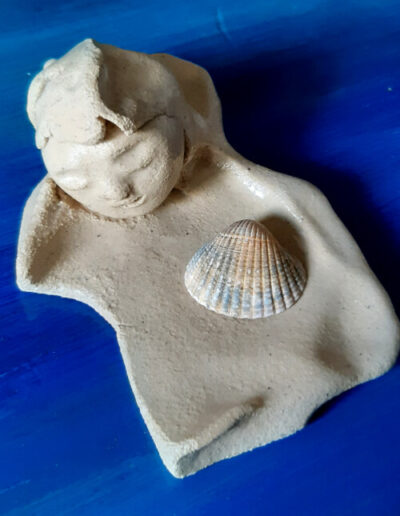 In der Draufsicht richtet sich der Blick auf einen flachen, hellen Teller, auf welchem eine Muschel liegt. Der Teller steht auf blauem Grund, hat Einkerbungen und geschwungene Rundungen. An einem Ende wird er begrenzt von einem still ruhenden Frauenkopf.