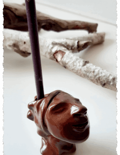 Ein individueller Kopf aus dunkelbraunem Ton, farblos glasiert, steht aufrecht auf glatter Fläche. Auf dem Kopf gibt es die Aussparung für eine Kerze, sodass ihm die Kerze zu entwachsen scheint.