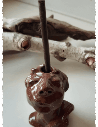 Ein individueller Kopf aus dunkelbraunem Ton, farblos glasiert, steht aufrecht auf glatter Fläche. Auf dem Kopf gibt es die Aussparung für eine Kerze, sodass ihm die Kerze zu entwachsen scheint.