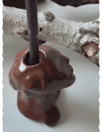 Ein individueller Kopf aus dunkelbraunem Ton, farblos glasiert, steht aufrecht auf glatter Fläche. Auf dem Kopf gibt es die Aussparung für eine Kerze, sodass ihm die Kerze zu entwachsen scheint.