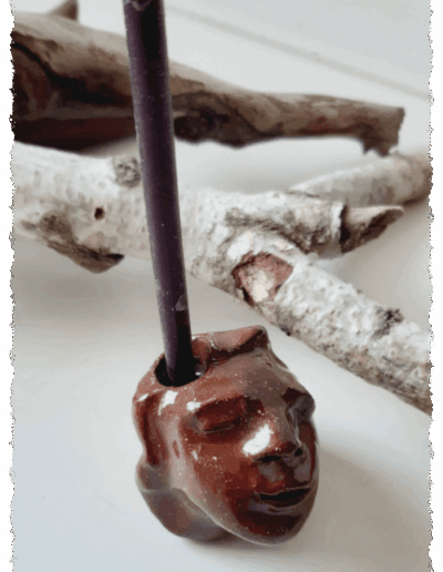 Ein individueller Kopf aus dunkelbraunem Ton, farblos glasiert, steht aufrecht auf glatter Fläche. Auf dem Kopf gibt es die Aussparung für eine Kerze, sodass ihm die Kerze zu entwachsen scheint.