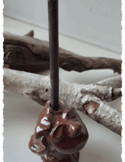 Ein individueller Kopf aus dunkelbraunem Ton, farblos glasiert, steht aufrecht auf glatter Fläche. Auf dem Kopf gibt es die Aussparung für eine Kerze, sodass ihm die Kerze zu entwachsen scheint.