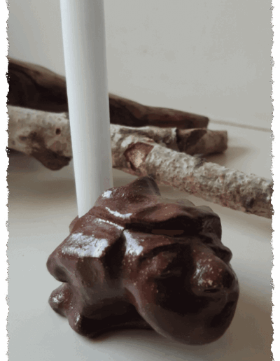 Ein individueller Kopf aus dunkelbraunem Ton, farblos glasiert, steht aufrecht auf glatter Fläche. Auf dem Kopf gibt es die Aussparung für eine Kerze, sodass ihm die Kerze zu entwachsen scheint.