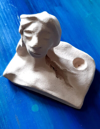 Ein sehr androgyner Kopf mit längerem Haar erhebt sich aus diesem kleinen, hellen, fast rechteckigen Tonteller. Er steht auf blauem Grund.Beginnend am Hals ist ein Riss sichtbar, der an eine Vulva erinnert. In einem Eck des Tellers befindet sich die Aussparung für eine kleine Kerze.