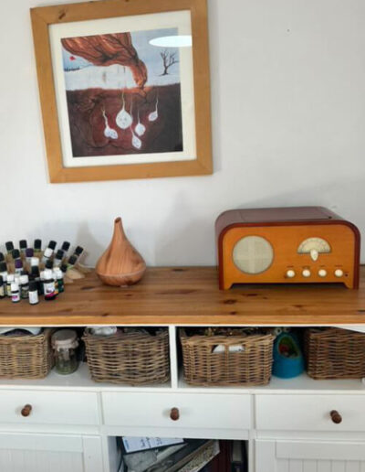 Die Malerei "Im Verborgenen" hängt in einem hellen Holzrahmen an einer weißen Wand in der Kindlein Komm Praxis von Simone Alwine Böhme. Unter ihr steht eine weiße Holzkommode, auf der Düfte, Öle und eine braunes Holzradio stehen.